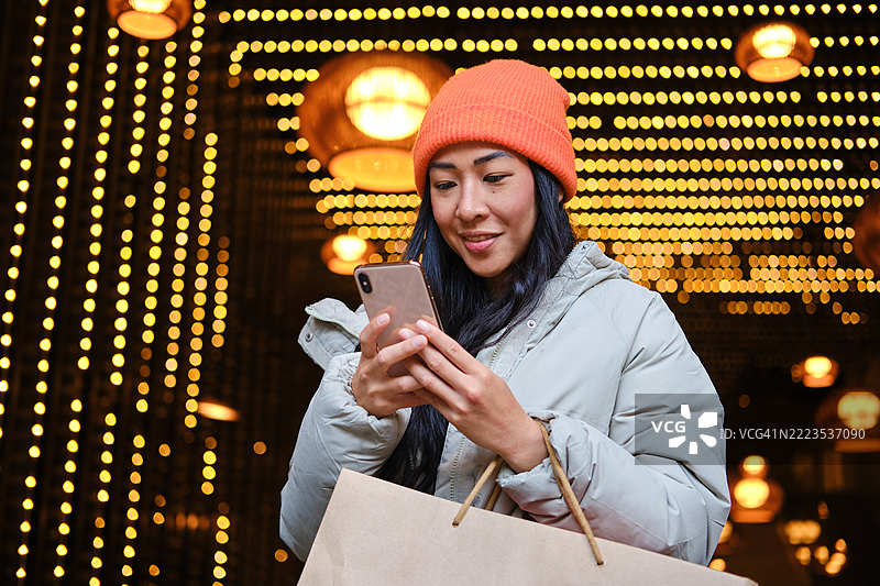 年轻的亚洲女性在圣诞购物时使用智能手机，背景灯光璀璨。图片素材