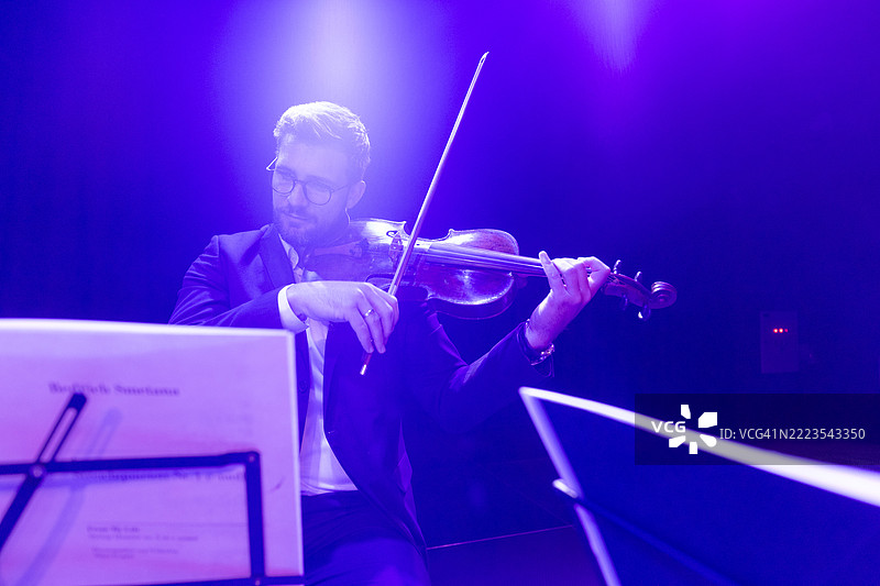 一位男性小提琴手在古典音乐会上演奏中提琴，专注地看着面前乐谱架上的乐谱。图片素材