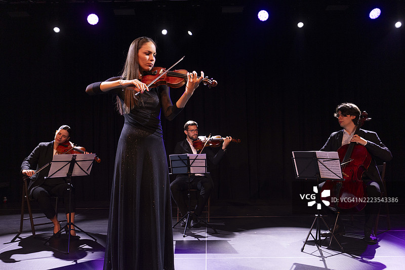 优雅的小提琴手穿着黑色优雅的裙子，站在弦乐四重奏的其他成员面前演奏小提琴。图片素材