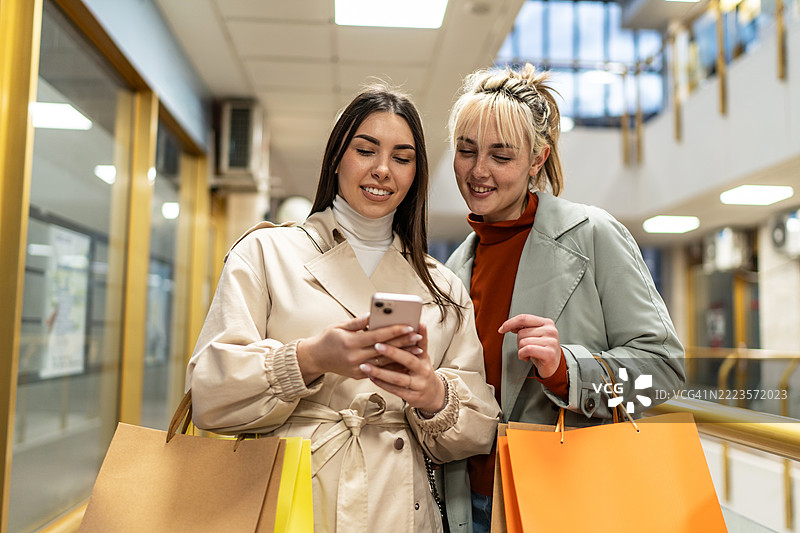 两位年轻女性在购物中心浏览手机在线优惠。图片素材