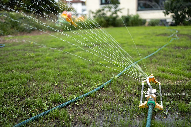 花园喷头在阳光明媚的日子里向郁郁葱葱的草坪喷洒水分，提供必要的水分补给。图片素材