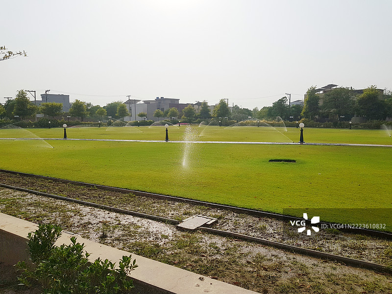 城市背景中建筑物后方的花园或地面上喷洒水的喷灌器。图片素材
