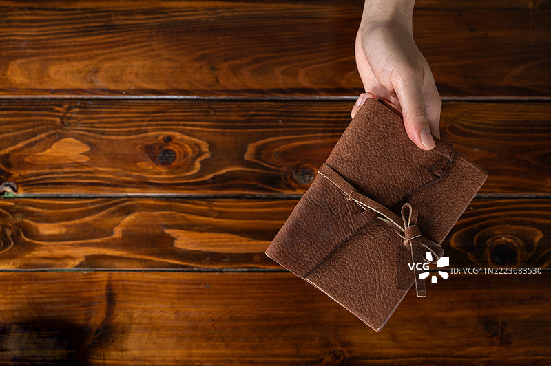高角度视图，手握皮革封面的书籍放在木桌上。图片素材