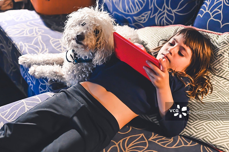 孩子在家庭客厅的沙发上放松，使用平板电脑，身边有她的宠物狗陪伴。图片素材