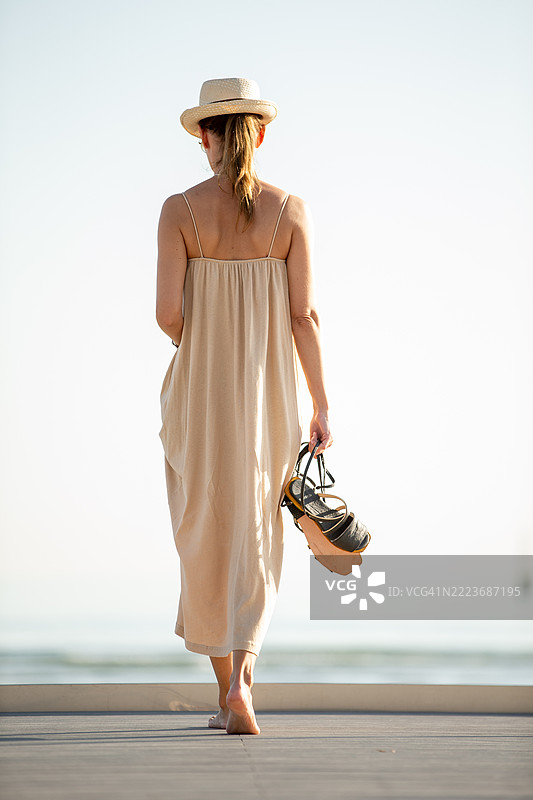 穿着长无袖裙子的女人在海边甲板上赤脚漫步，享受着悠闲时光。图片素材