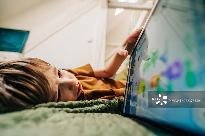 无聊的男孩躺在家里的沙发上使用平板电脑。图片素材