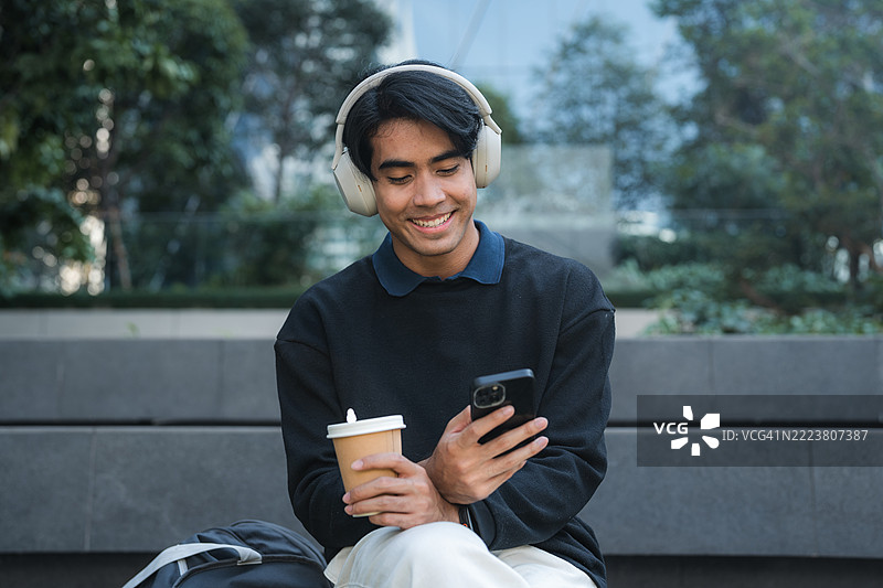年轻男子在 sidewalk 旁放松，戴着耳机听音乐，同时滑动手机。图片素材