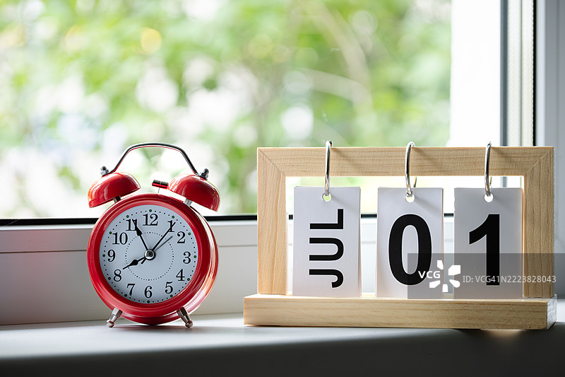 红色闹钟显示早上8点，窗台上的木质桌面日历显示日期为7月1日，背景是窗户。图片素材