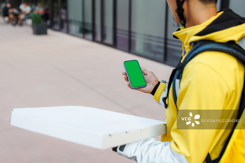 穿着黄色夹克的送餐员手持披萨盒和带有绿色屏幕的手机图片素材