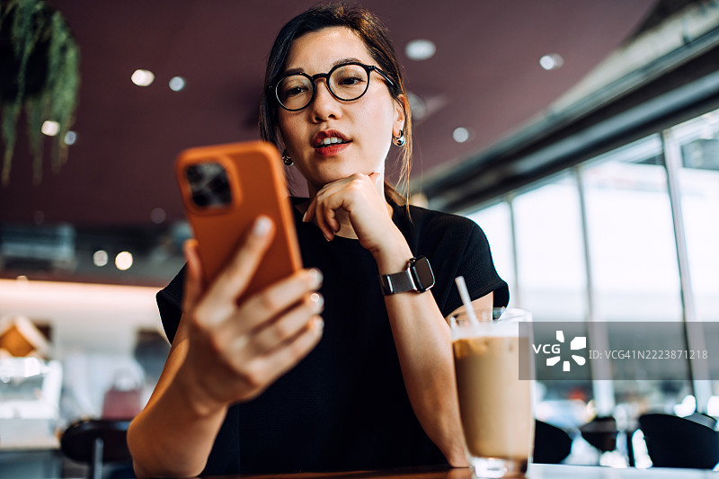 自信时尚的亚洲女商人在咖啡馆喝咖啡并使用智能手机。管理个人财务和在线银行业务。查看投资和股票市场交易，使用手机制定财务计划。财富管理和在线银行业务。图片素材
