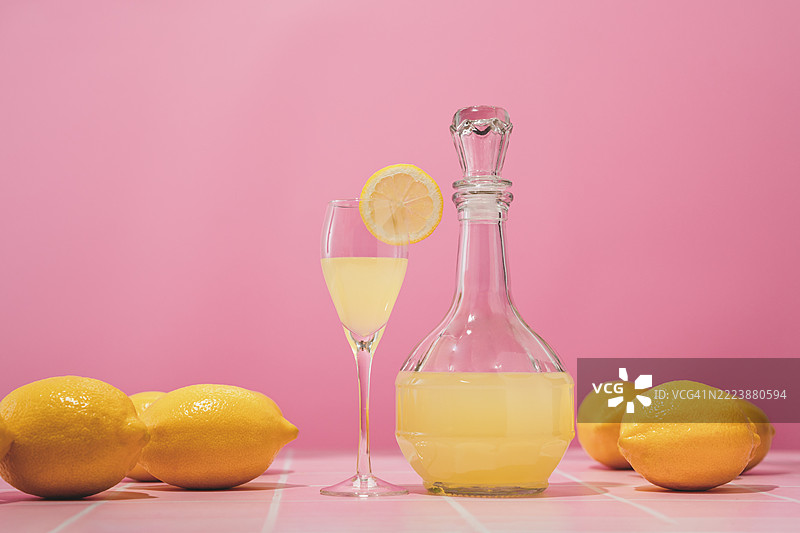 柠檬酒。几颗新鲜的完整柠檬散落在粉色桌子上，背景也是粉色，留有空白空间。夏季花园，自制柠檬利口酒装在玻璃壶中。图片素材
