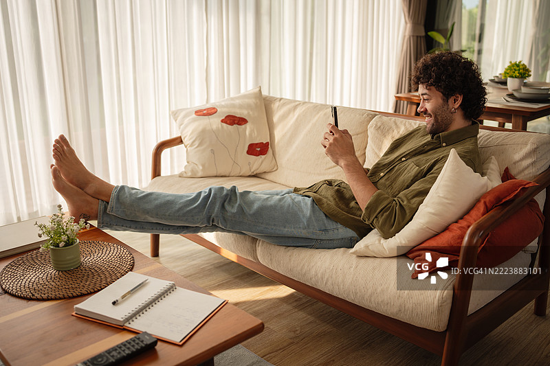 年轻男子坐在沙发上放松，手里拿着手机图片素材