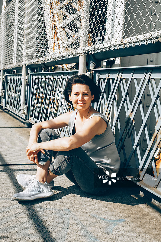 年轻女性坐在阳光下的人行天桥上，放松的姿势，面向镜头。图片素材