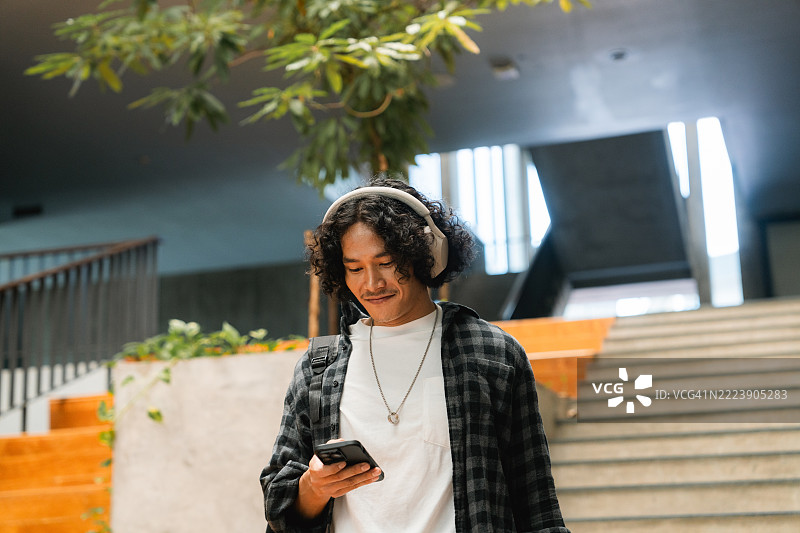 微笑的大学生在校园里走着，手持数字平板，戴着耳机听音乐。图片素材