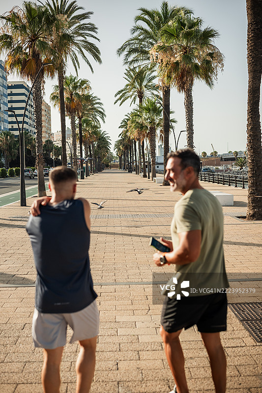 两名男子在棕榈树成行的海滨长廊上跑步后拉伸身体图片素材