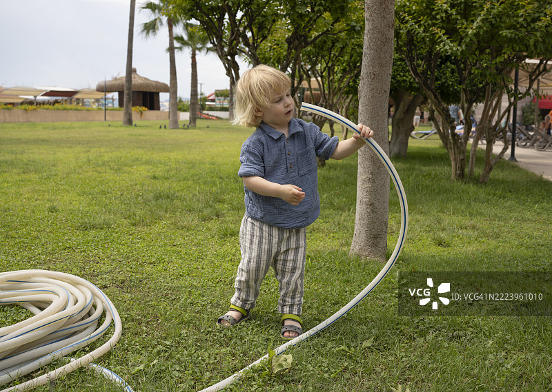 好奇的金发幼儿在公园般的环境中，草坪上玩着一根大水管。这是一个户外探索和童年游戏的自然瞬间。图片素材