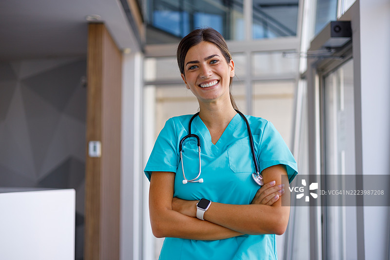 自信的女性医生微笑着，双臂交叉，佩戴听诊器和智能手表，站在现代医疗环境中。图片素材