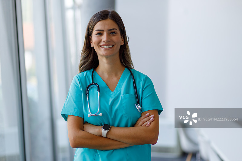 一位自信的年轻女医生微笑着，双臂交叉，站在现代医院走廊中。图片素材