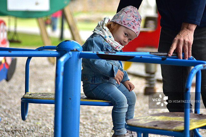 可爱的幼儿女孩穿着牛仔服,坐在蓝色游乐场旋转木马上,顽皮地歪着头,成人的手在一旁提供支持。在公园享受户外乐趣和亲密时光,立陶宛,考纳斯地区。图片素材