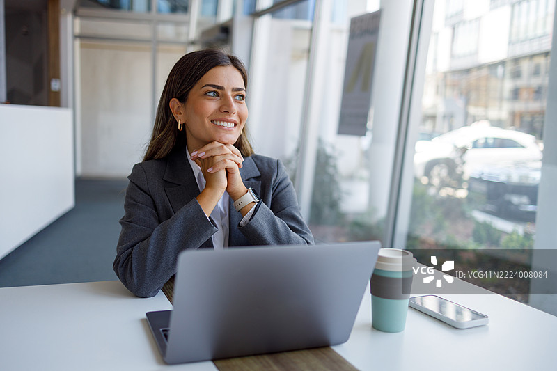 年轻女商人在现代办公室的桌子前坐着，面带微笑，目光投向一旁，享受着她的工作。图片素材
