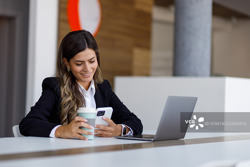 微笑的女商人在现代办公室的桌子上使用智能手机并喝咖啡，同时旁边有一台笔记本电脑。图片素材