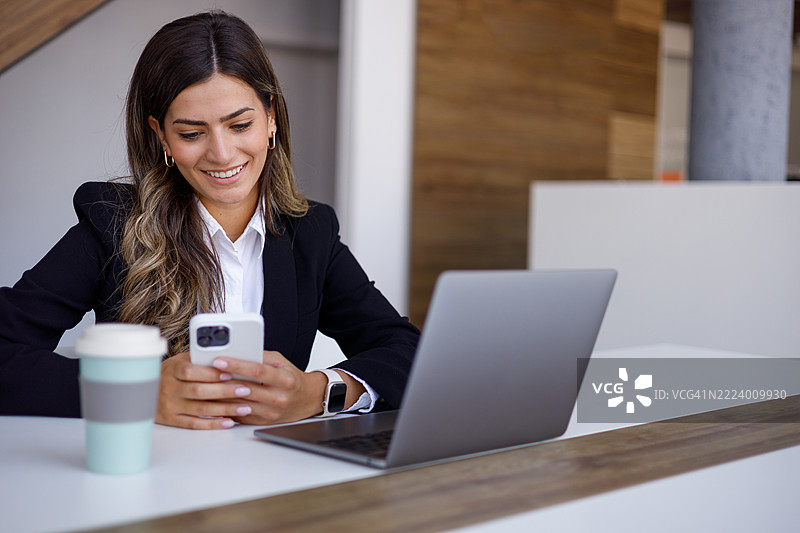 商务女性在现代办公室的桌子上坐着，使用智能手机并微笑，同时旁边有笔记本电脑和咖啡。图片素材
