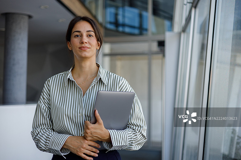 专业女商人握着笔记本电脑，站在现代企业办公空间的玻璃窗旁，自信地凝视着观众。图片素材