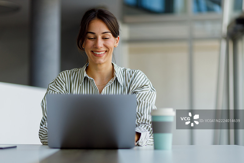自信微笑的职业女性在现代工作空间中使用笔记本电脑打字。图片素材