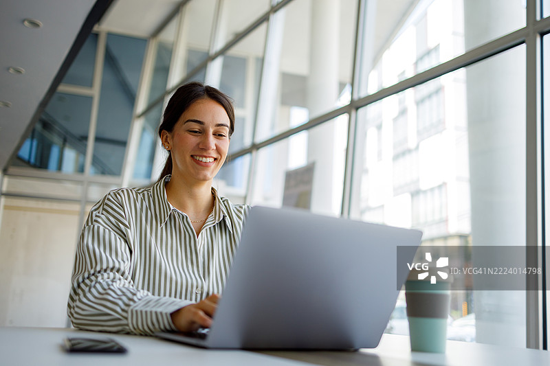 年轻女商人在明亮现代的办公室里微笑着使用笔记本电脑，办公室有大窗户和充满活力的氛围。图片素材