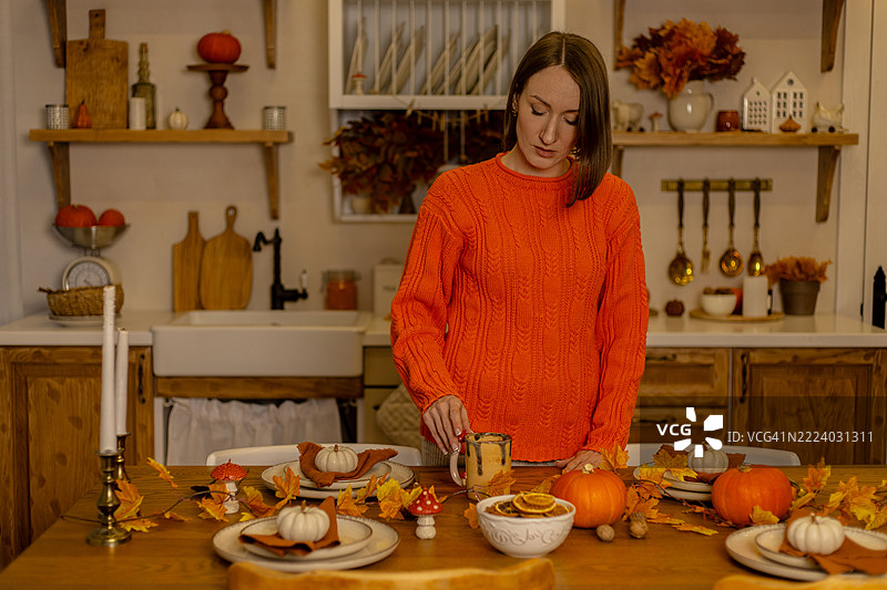 穿着橙色毛衣的年轻女性在一个节日气氛浓厚的秋季乡村餐桌上拿起一个杯子。图片素材