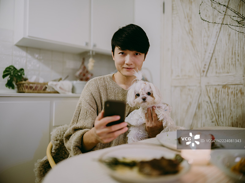 年轻帅气的亚洲男子在餐厅里抱着小狗并使用智能手机图片素材