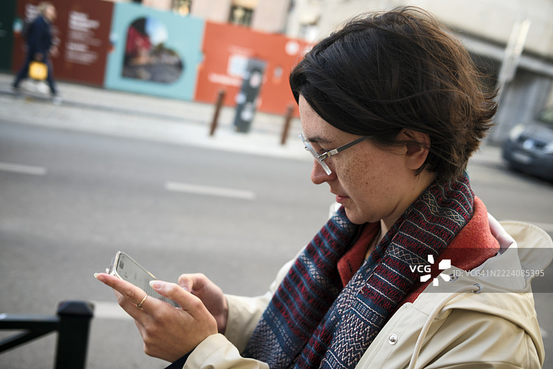 戴眼镜的女性在城市街道上浏览手机，穿着图案围巾和米色外套。图片素材
