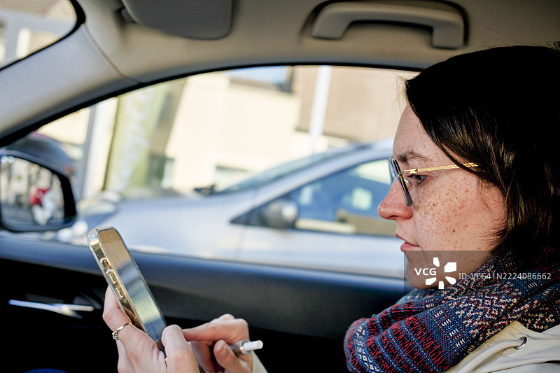 阳光明媚的日子里，法国勃艮第，一位戴着眼镜和围巾的女性在车内使用智能手机。图片素材