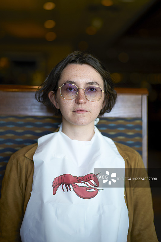 年轻女性戴着眼镜，穿着印有红色龙虾图案的白色围裙，坐在室内，背景模糊。美国棕榈泉图片素材