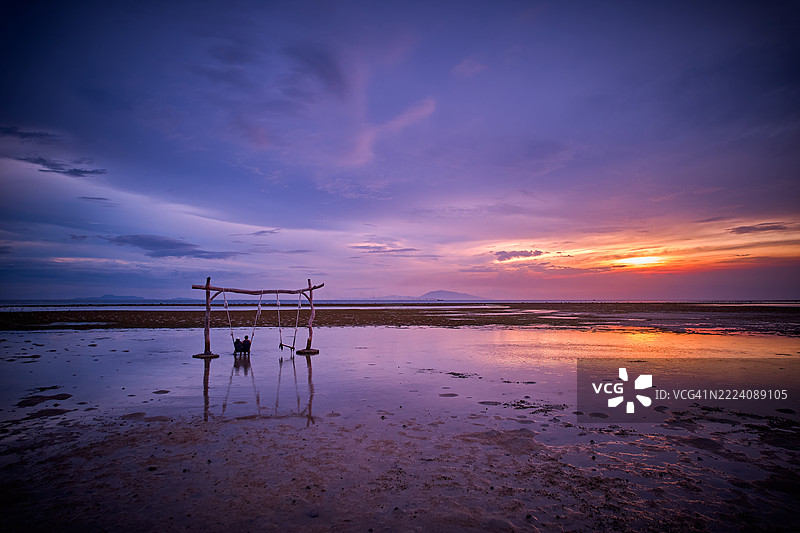 一个木制秋千架矗立在日落时分海滩的浅水中。图片素材