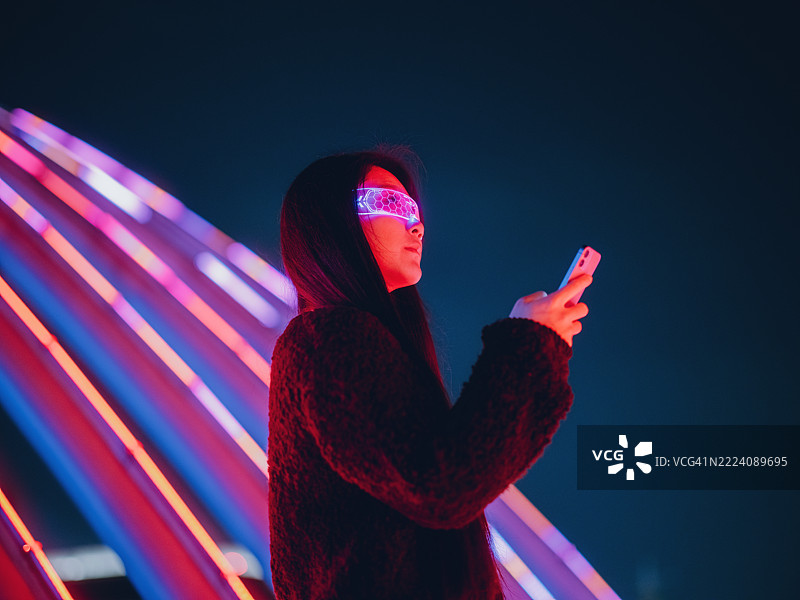 年轻的亚洲女商人在霓虹灯闪烁的街道上站着。佩戴着发光眼镜，使用智能手机。人工智能与网络安全概念图片素材