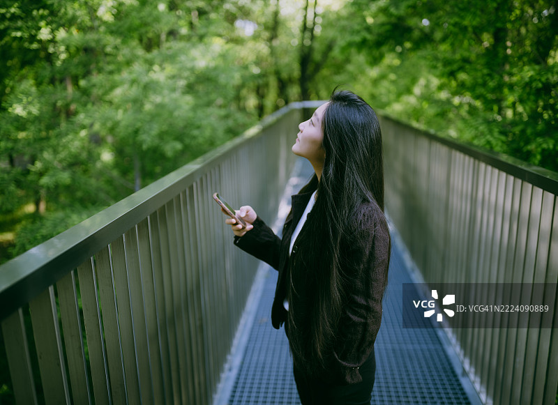 年轻的亚洲女商人在森林中站着，使用智能手机。呼吸新鲜空气，亲近自然。图片素材