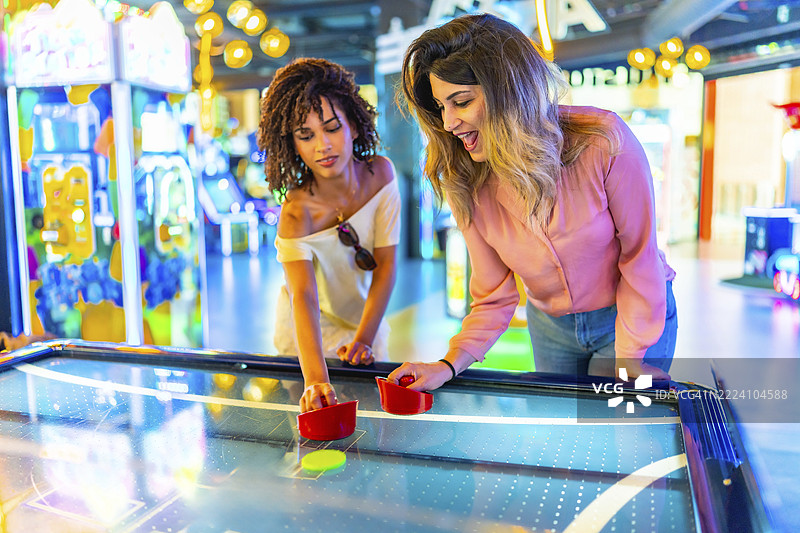两位年轻女性在一个充满活力的游戏厅里愉快地进行一场友好的空气曲棍球比赛，展现了室内娱乐的乐趣与兴奋。图片素材