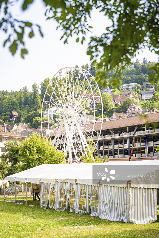 大型白色帐篷后面的摩天轮,周围是村庄房屋和绿色山丘,摩天轮是为了庆祝德国黑森林卡尔夫镇950周年而建造的。图片素材