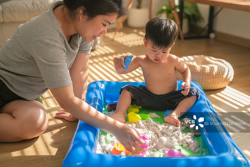 亚洲母亲和她的幼儿儿子在家中享受快乐的周末，一起玩疏水沙。小男孩在家里的沙箱中进行感官游戏，帮助发展精细运动技能。图片素材