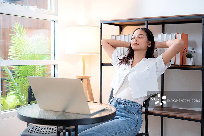 年轻的亚洲女性在舒适的家中放松，远程工作后使用笔记本电脑，享受安静的空间，关注自由职业和身心健康，生活方式与工作空间，自由职业者在家办公室的桌子旁休息。图片素材