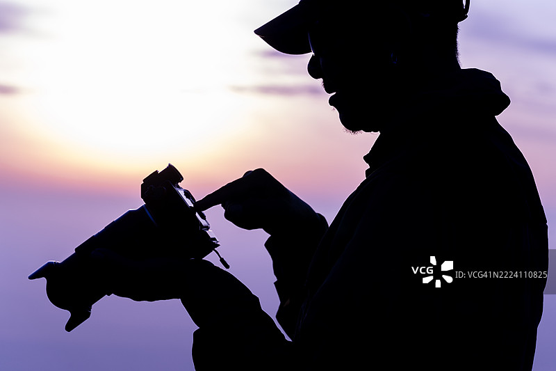日出时分，男子在户外摄影冒险中审视相机的剪影，激发灵感与创造力。图片素材