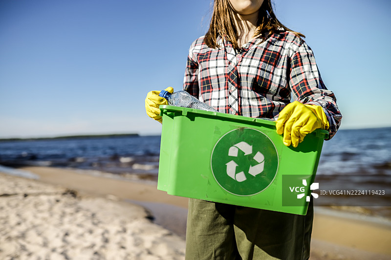 河流清理行动：一名未知的青少年在河边分类丢弃的瓶子，将它们放入指定用于二次处理的绿色垃圾箱中。图片素材