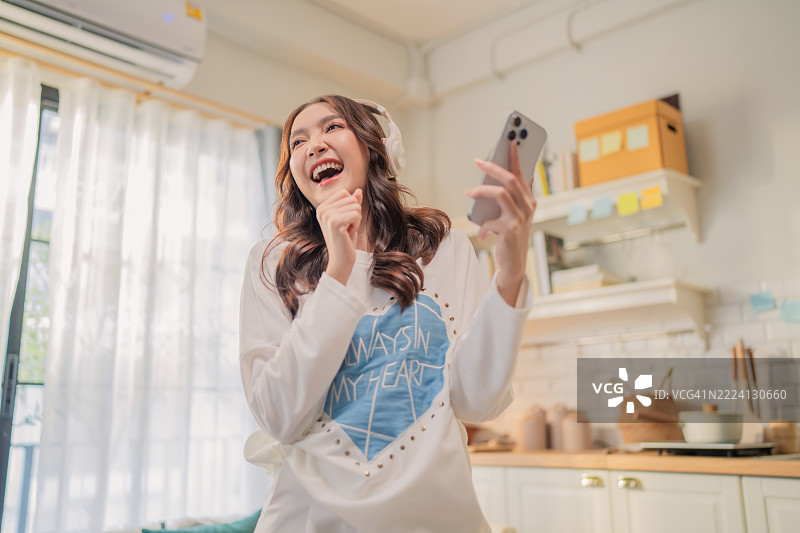 美丽的亚洲女性站在舒适的客厅里，使用智能手机，戴着耳机听音乐。她脸上洋溢着快乐，自由地跳舞并跟着唱歌。图片素材