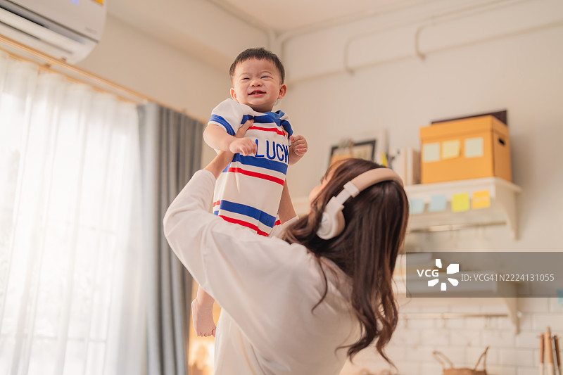 年轻的亚洲母亲在温馨的厨房里将婴儿男孩高高举起，愉快地玩耍。这个家庭生活场景捕捉了母子之间温暖的亲密关系和快乐的育儿时刻——一种快乐的生活与爱的概念。图片素材