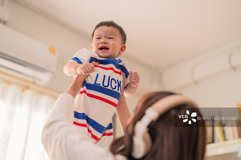 年轻的亚洲母亲在温馨的厨房里把婴儿男孩举起来，愉快地玩耍。这个家庭生活场景捕捉了母子之间温暖的亲密关系和快乐的育儿时刻——一种快乐的生活与爱的概念。图片素材