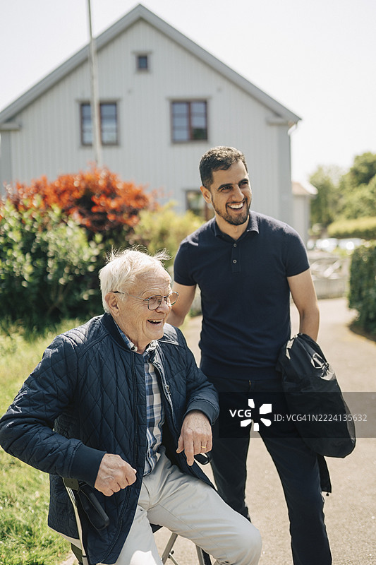 快乐的男性护理助理与坐在助行器上的老人在阳光明媚的公园里。图片素材