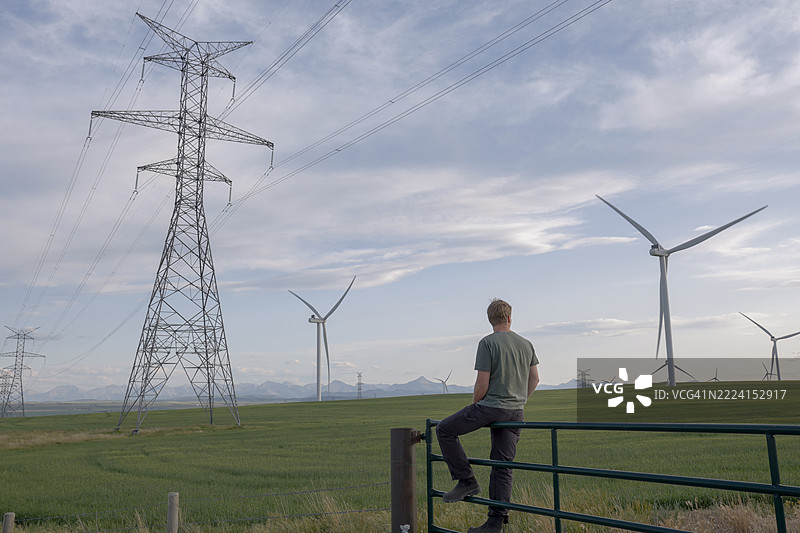 年轻男子坐在围栏上，凝视着风力发电机。图片素材