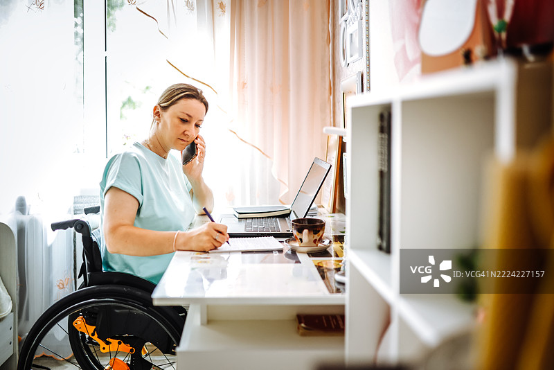 坐在轮椅上的女性在家中使用笔记本电脑，同时打电话。图片素材