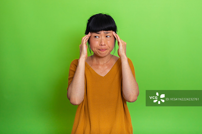 一位女性双手抱头，感到偏头痛，背景为绿色。图片素材